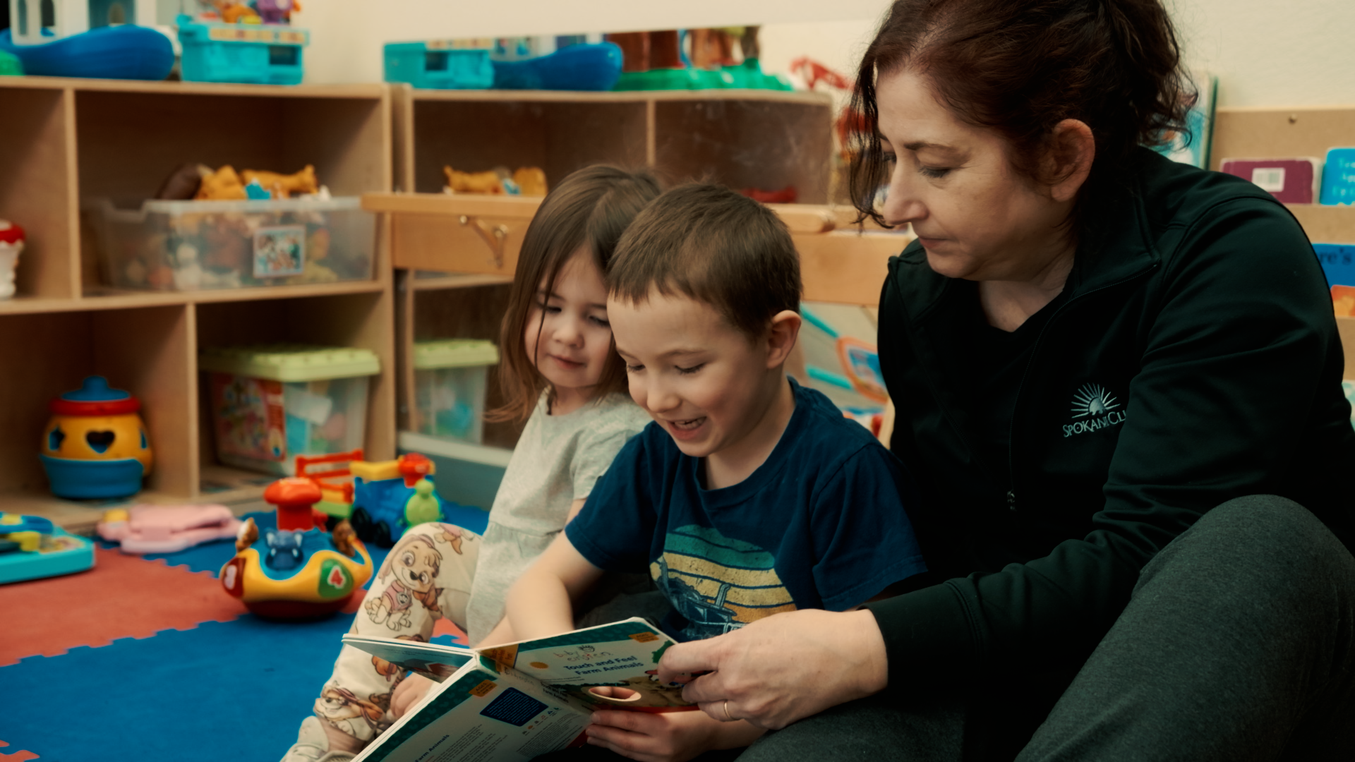 Tracey from the Spokane Club childcare team sits with a couple young kids and reads them a story. The Spokane Club offers 3 hours of complimentary childcare every day for family memberships.